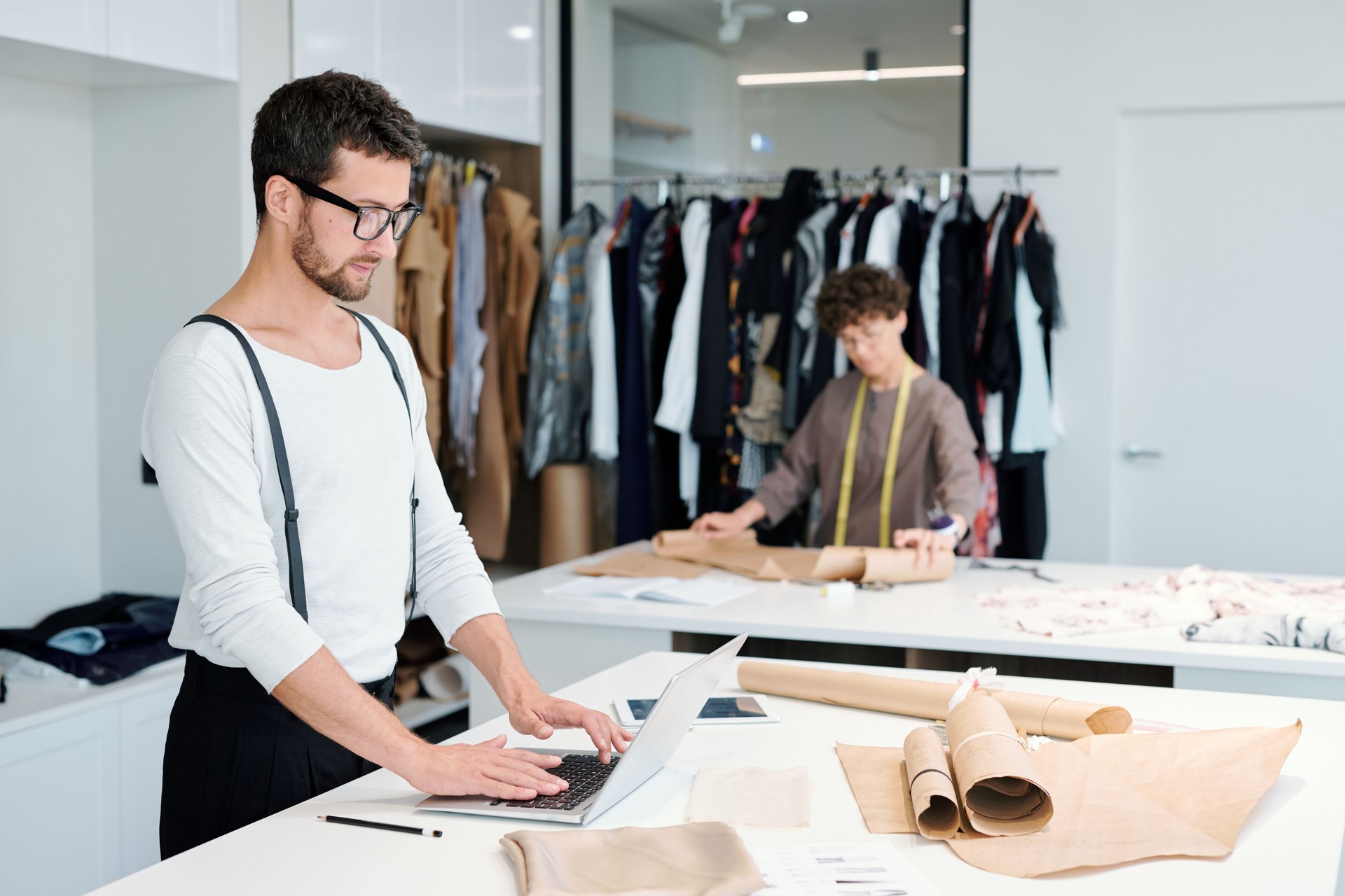 Young tailor in casualwear standing by desk in front of laptop