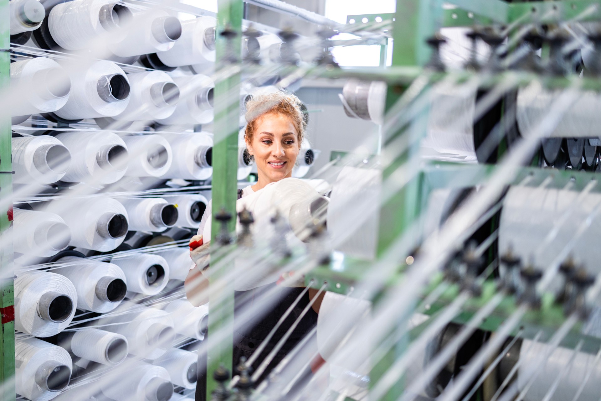 Female textile factory worker changing thread spool on industrial knitting machine.