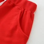Close-up of baby red shorts detail, featuring a thick ribbed elastic waistband, precision-stitched side pocket, and premium vibrant cotton fabric. High-quality infant apparel construction from a professional clothing manufacturer.